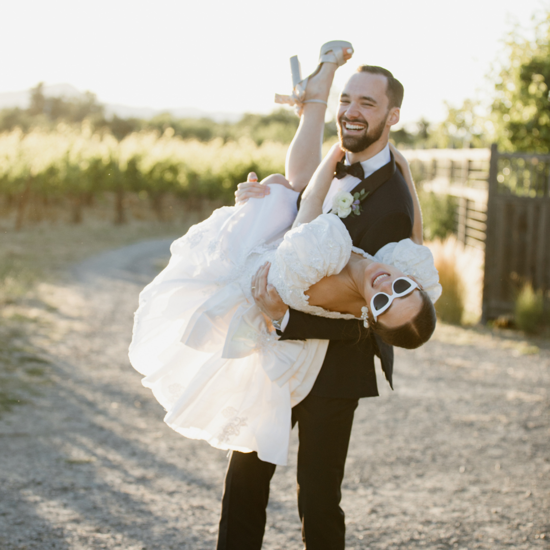 Couple laughing as the groom carries his bride down a Napa vineyard road